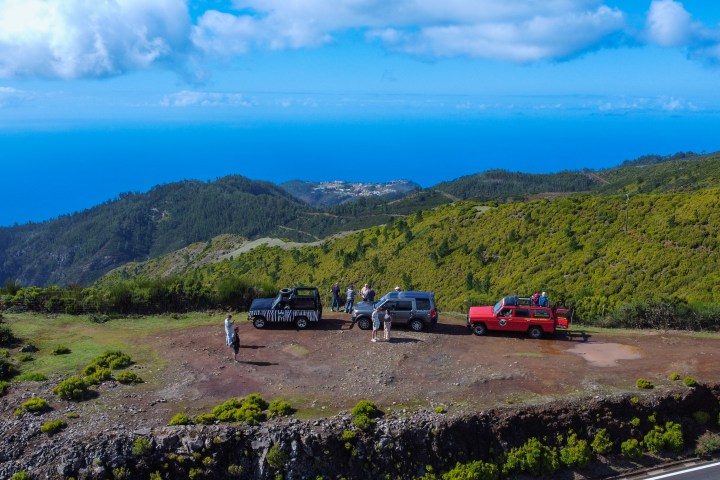 Three parked SUVs on a scenic hilltop with people enjoying the view of the ocean and lush green hills.