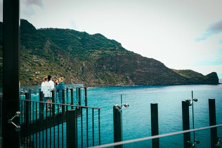 People standing on a glass balcony overlooking a large body of water and mountains.