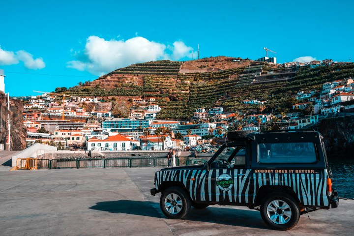 Zebra-striped Jeep parked on dock with hillside town and blue sky in background.