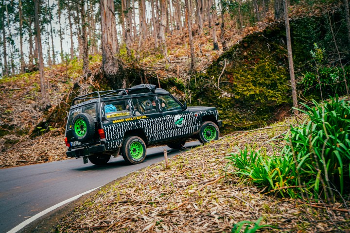 A black SUV with neon green wheels drives on a forest road surrounded by trees.