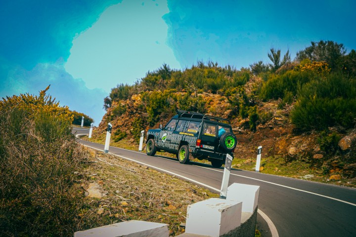 Car with green wheels drives on winding mountain road under blue sky.