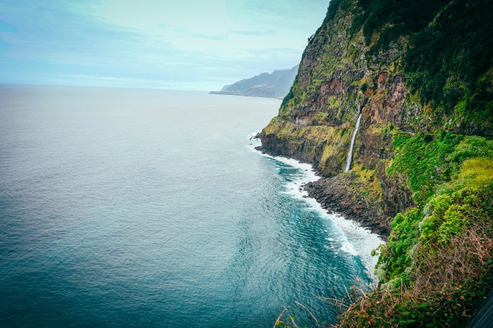 Seaside cliffs with a waterfall flowing into the ocean under a cloudy sky.