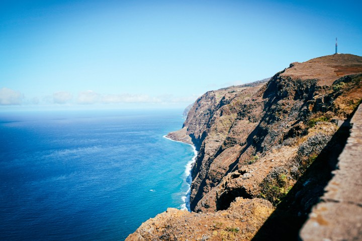 Cliffside view of ocean with clear blue sky and waves reaching the rocky shore.