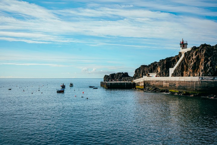 Coastal scene with a lighthouse on rocky cliffs and boats in the water at Madeira island