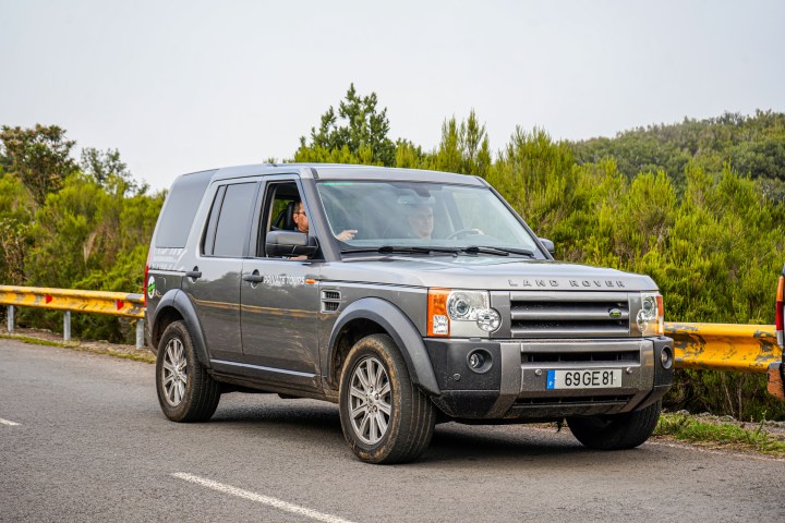 Gray Land Rover on road with trees and yellow barrier in background.