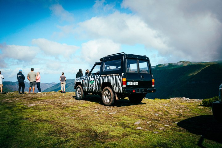 Off-road vehicle and people on grassy mountain lookout with cloudy sky.