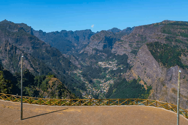 Mountainous landscape with a fenced viewpoint, overlooking valleys and distant village under clear blue sky.