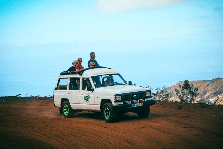 White off-road vehicle with people on roof, driving on a dirt road with ocean in the background.